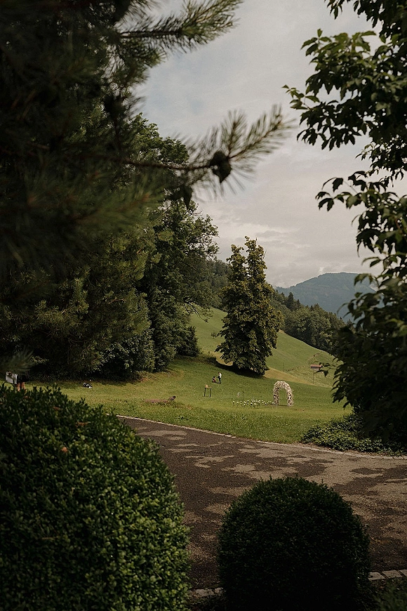 Outdoor ceremony setup with a floral arch and rows of chairs on a grassy hillside, with trees and mountains under a cloudy sky