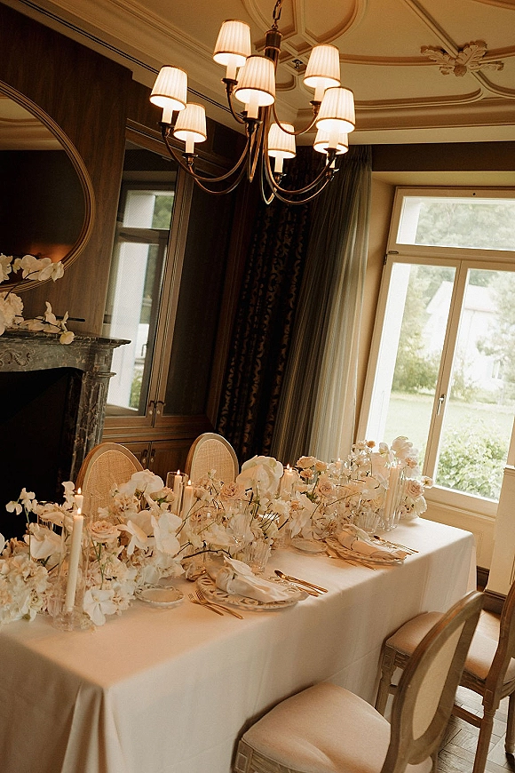 Reception tablescape with head table decor, white roses and hydrangea centerpiece, taper candles and glassware beneath chandelier by fireplace