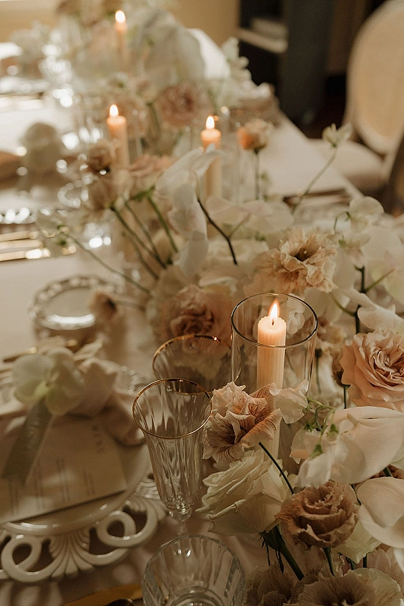 Reception tablescape with a wedding table centerpiece of blush roses, white florals, and taper candles in glass hurricane holders on white linens indoors