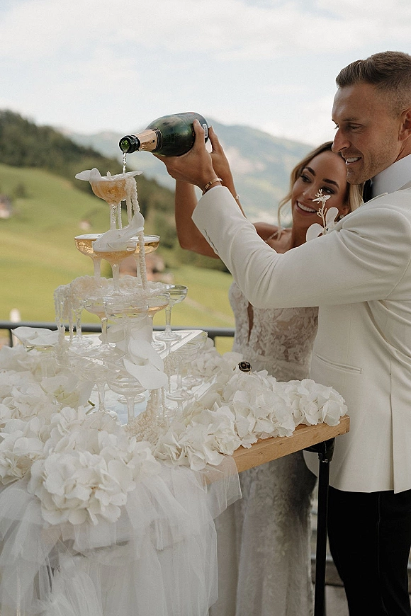 Champagne tower toast as bride and groom pour into coupe glasses with white floral garland on a terrace overlooking mountains under clouds