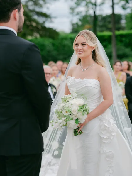 Ceremony moment during outdoor wedding vows, bride facing groom and holding a white rose bouquet in a garden with guests seated.