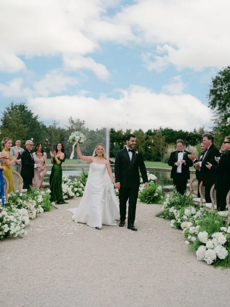 Ceremony recessional captures a just married moment as the bride and groom walk the gravel path by a fountain, showered with rose petals.