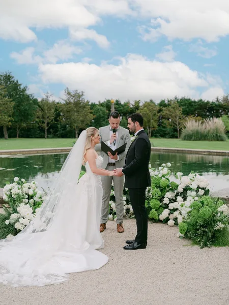 Ceremony moment as the couple, hands clasped during the exchange of vows, her long veil trailing on the lawn by a pond and garden trees.
