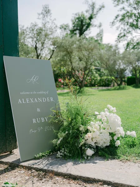 Wedding welcome sign doubles as a ceremony entrance sign, trimmed with white florals beside a green post on a stone path under garden trees.