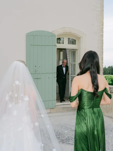 Father daughter first look during a dad sees bride moment by an arched doorway with green shutters, her bridal veil trailing across the stone patio.