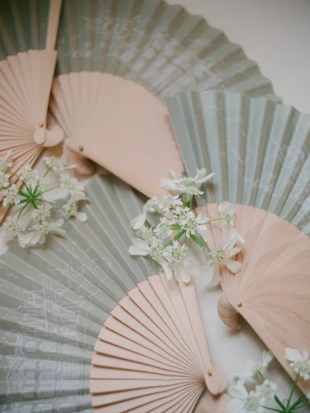 Wedding fans arranged in a flatlay: folding hand fans and paper hand fans with wood handles, accented by white flowers on soft linen.