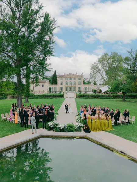 Wedding recessional with a couple walking down the aisle past aisle flowers on a manicured lawn, mansion facade and reflecting pool beyond.
