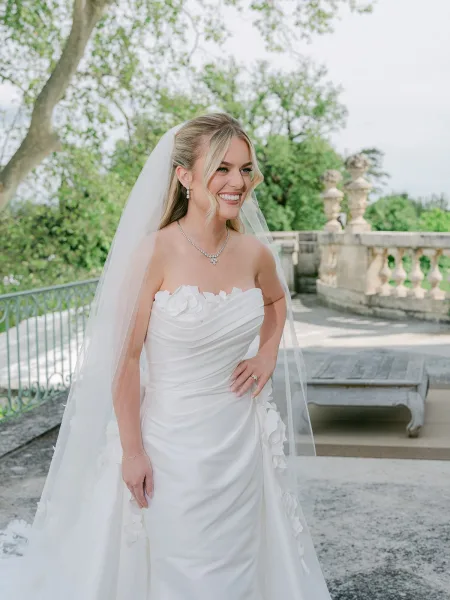 Bridal portrait of a bride with veil on an outdoor terrace by a stone balustrade, garden trees behind, face-framing curls and a warm smile.