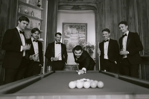 Groom with groomsmen play pool around a billiards table in a wood-paneled lounge, laughing as the groom lines up a shot.