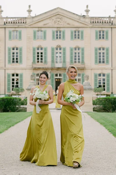 Bridesmaid dresses in chartreuse satin sway as the bridal party walks with white bouquets past a stone estate facade and manicured hedges.
