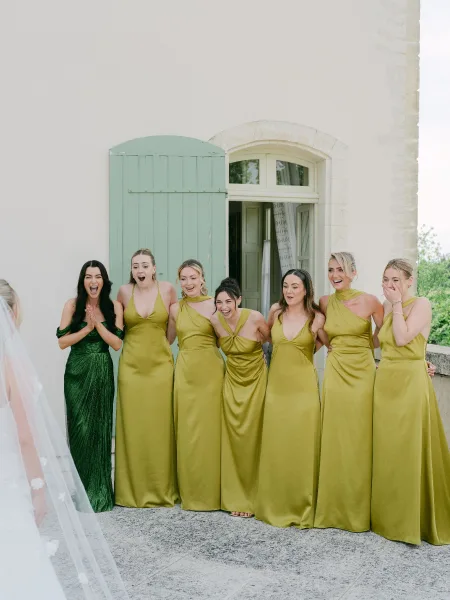Bridesmaids first look during the bride reveal to bridesmaids, beaming as her airy bridal veil catches light by a mint green shutter.