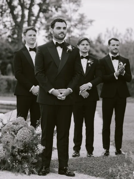 Ceremony moment with the groom waiting at the altar in a black bow tie, framed by garden trees along the lawn pathway.