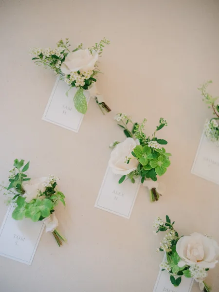 Wedding boutonniere, a white rose boutonniere with a name tag, neatly arranged on a neutral beige fabric backdrop.