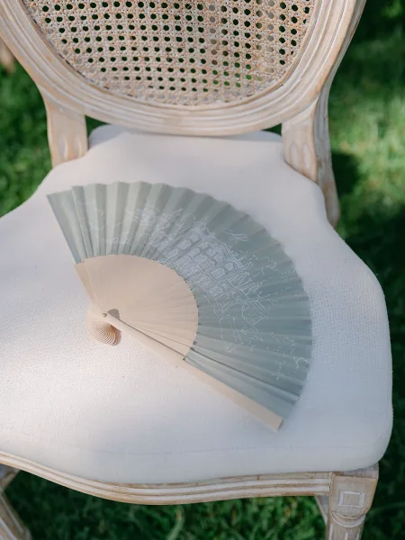Wedding fan favor, a hand fan for wedding guests, rests on a cane-back chair atop a grass lawn during an outdoor ceremony.