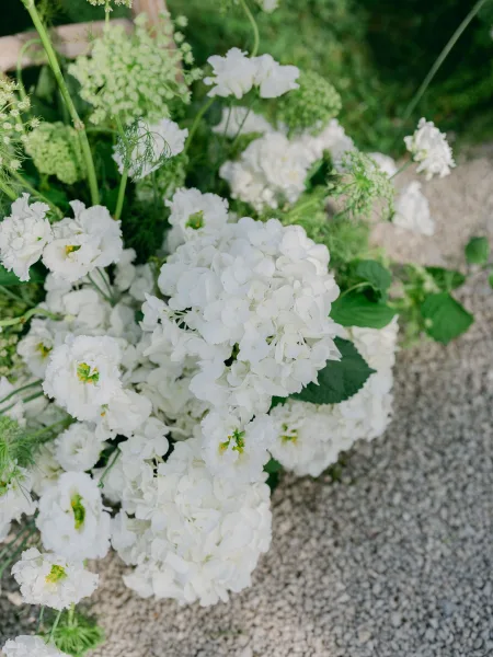 Wedding floral arrangement of white and green florals featuring white hydrangeas, set along a gravel path amid lush garden greenery.