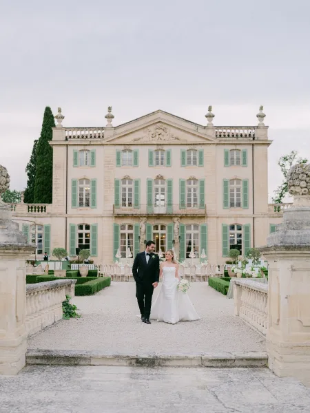 Couple portrait of the bride and groom walking hand in hand along the chateau's gravel path, her white bouquet bright against an overcast sky.