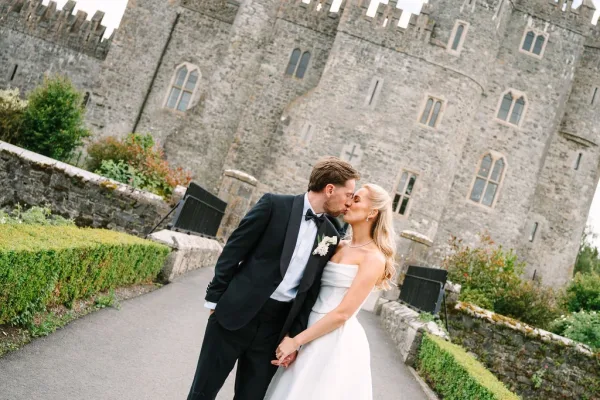 Wedding kiss portrait of a bride and groom holding hands, the groom's white rose boutonniere standing out on a stone castle walkway.