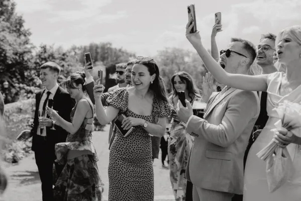 Wedding guest reactions with guests cheering, some lifting smartphones, in a leafy garden as friends and family beam along the pathway.