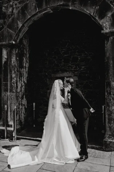 Wedding kiss portrait of a bride and groom under a stone archway, her lace sleeves catching light as they embrace on the worn stone floor