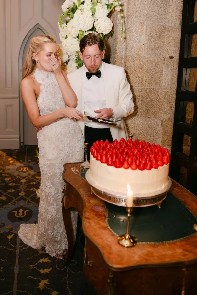Wedding cake cutting as the couple slices a strawberry wedding cake on an antique table beside a stone wall with arched door.