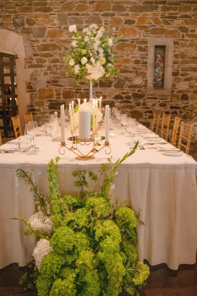 Reception table decor for a long banquet table setup with a gold candelabra, set against a stone wall and arched doorway.