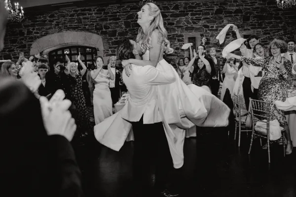 Reception entrance for a newlywed grand entrance, groom lifting the bride as guests wave napkins by an arched stone window in the banquet hall.