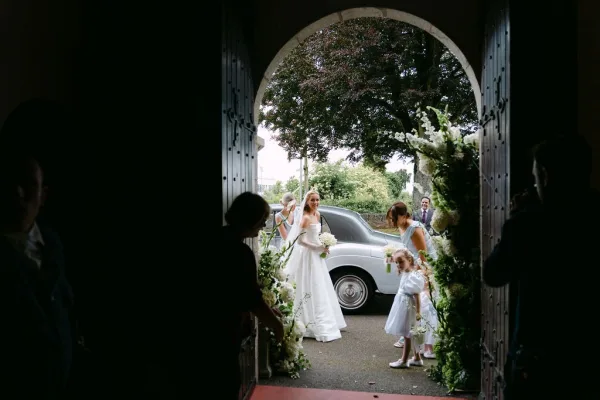 Bride arrival at the church door, holding a white bouquet beside the arched doorway, ready to step inside as the church doors frame the scene.