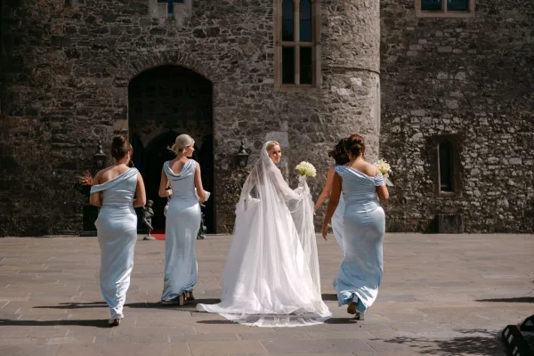 Bride and bridesmaids walking as a bridal party in a stone courtyard by an arched doorway, the bride wearing a long veil.