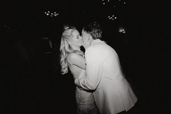 Wedding kiss portrait capturing a dance floor kiss as the bride and groom embrace, the groom in a tuxedo jacket, on a dark reception dance floor.