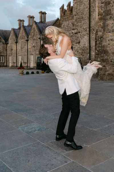 Groom lifting bride in a joyful couple lift pose, her lace bridal gown floating, in a stone courtyard by a historic building.