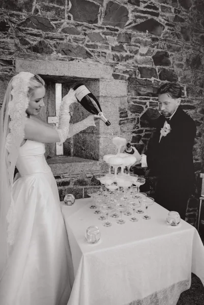 Champagne tower pour as a bride pouring champagne joins her groom, cascading into coupe glasses by a stone wall, lace gloves catching light.