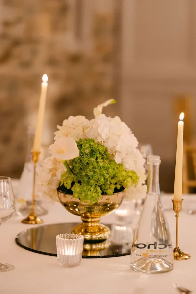 Wedding centerpiece, a gold compote centerpiece brimming with white hydrangeas, set before a stone wall at an indoor reception.