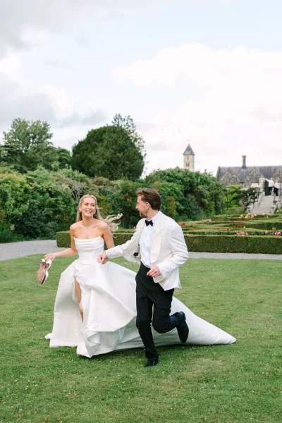 Couple portrait of a bride and groom running hand in hand across a manicured lawn, her pearl necklace catching light by a stone building.