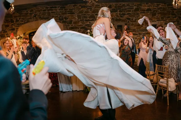 Reception dance moment with a wedding dance lift as the groom raises the bride, guests cheer and wave napkins beside a stone wall and arched window.
