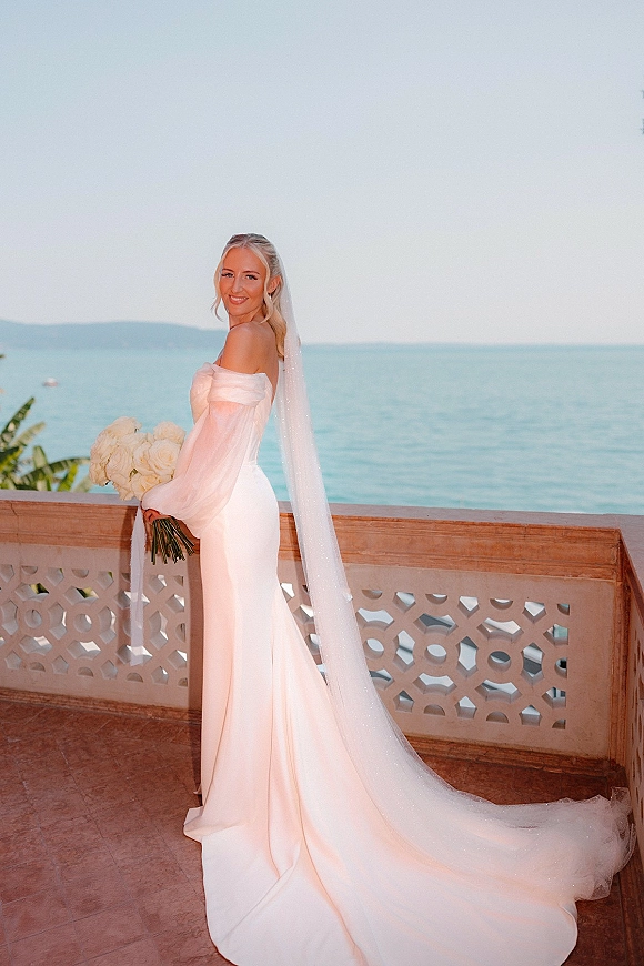Bridal portrait of a bride holding bouquet of white roses, wearing an off-the-shoulder dress and long veil on an oceanfront terrace
