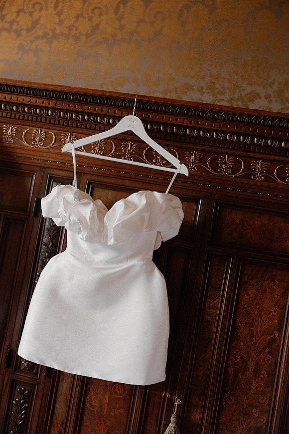 Wedding dress in a short wedding dress style hangs on a white dress hanger, puff sleeves framed by a carved wood cabinet and wallpaper