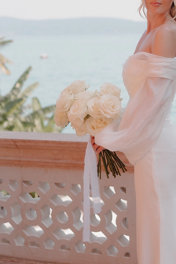 Bridal portrait of a bride holding bouquet of white roses with trailing ribbon, in an off shoulder dress on a balcony with ocean and palm leaves