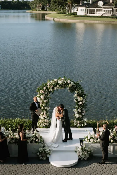 Ceremony kiss under a round white rose and greenery floral arch, bride in strapless gown and veil, lakeside lawn backdrop
