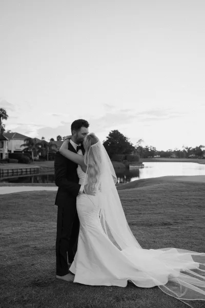 Wedding kiss portrait of bride and groom embracing, her long veil and dress train flowing beside a lakeside lawn with trees and homes
