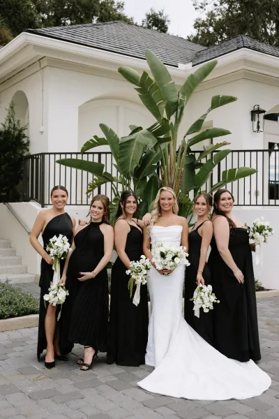 Bridesmaid group photo with bride and bridesmaids in black bridesmaid dresses holding white bouquets on steps by a white stucco building.