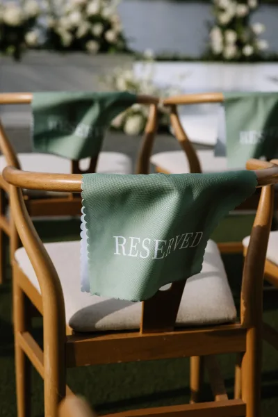 Reserved chair sign in green cloth with white lettering draped over wooden wedding chairs near a floral ceremony stage on grass lawn