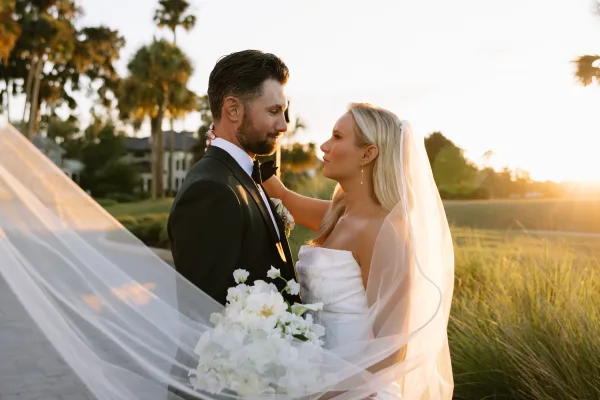 Couple portrait of bride and groom at sunset, forehead to forehead as her veil blows, holding a white bouquet with palm trees behind them