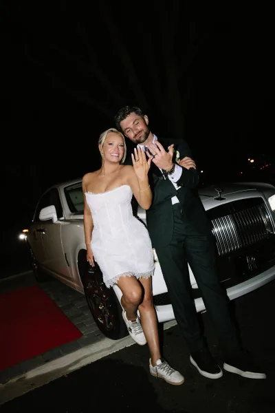 Couple portrait of newlyweds showing wedding rings while leaning on a luxury car at night beside a red carpet runner and street lights