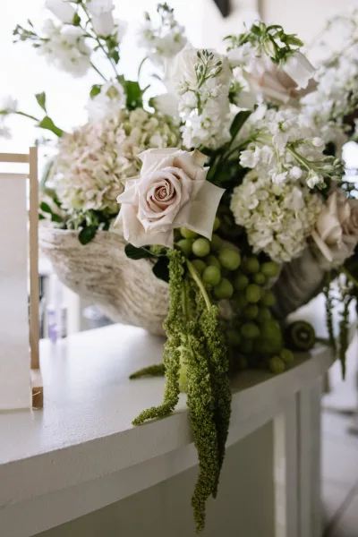 Wedding floral centerpiece in a stone compote bowl with white roses, hydrangea, stock, greenery, hanging amaranthus, and green grapes in bright window light