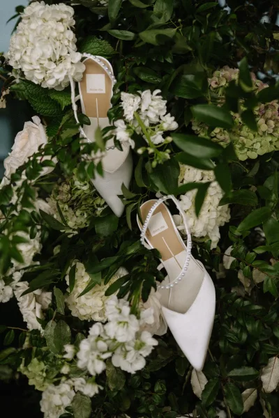 Bridal shoes, pearl wedding heels with pointed-toe design and pearl ankle straps nestled among flowers and greenery in a flat lay