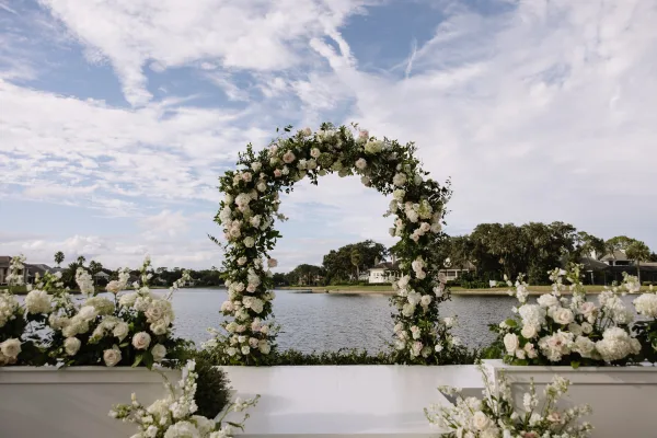 Wedding ceremony arch with a round wedding arch of white and blush roses, hydrangeas, and greenery on a lakefront lawn under clouds