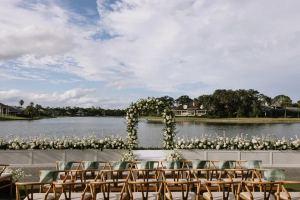 Ceremony setup for a waterfront wedding ceremony with a floral arch, white and green arrangements, wooden chairs, and a lake backdrop under clouds