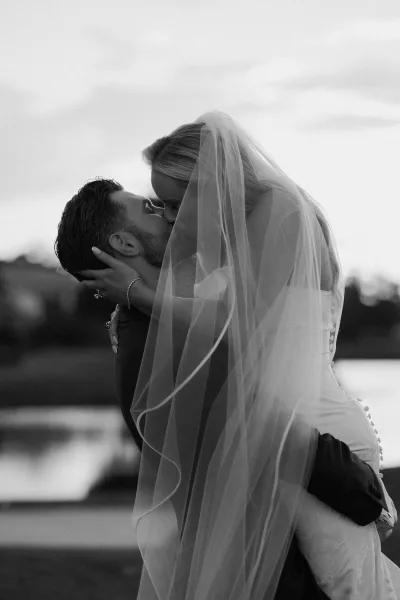 Wedding kiss portrait of bride and groom kissing in a dip, veil draped over them by a lakeside landscape with trees and sky