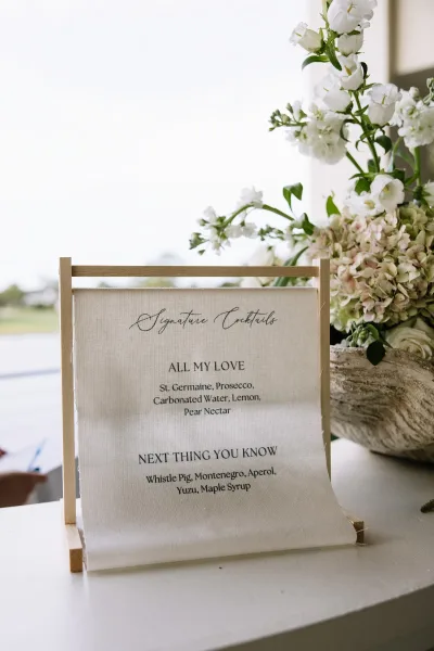 Signature cocktails sign with calligraphy wedding bar menu sign on linen in a wooden stand, paired with white flowers and greenery on an outdoor table