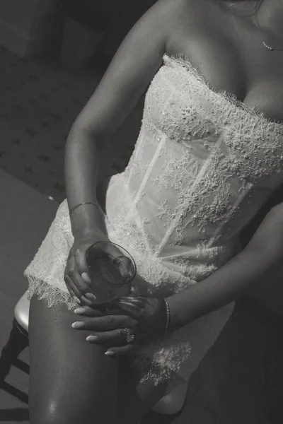 Bridal lingerie in a lace bridal bodysuit, seated on an upholstered chair, showing engagement ring and manicure while holding a glass tumbler.